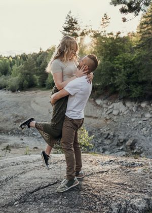 Mann hebt seine Freundin in die Höhe auf einem Felsen stehend zum Sonnenuntergang am Forstsee